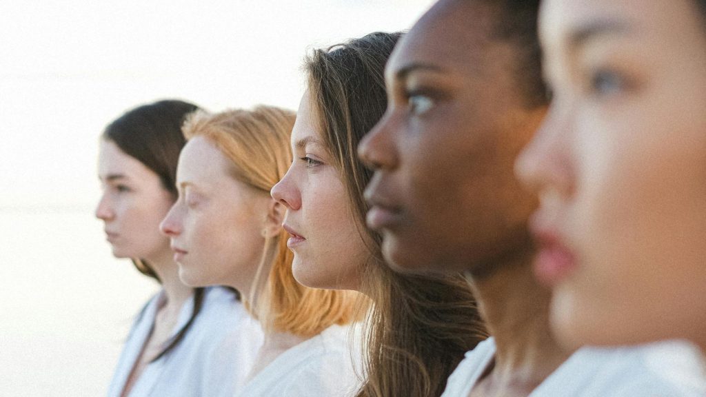 Five women of different ethnicities standing together by the sea, embracing diversity.
